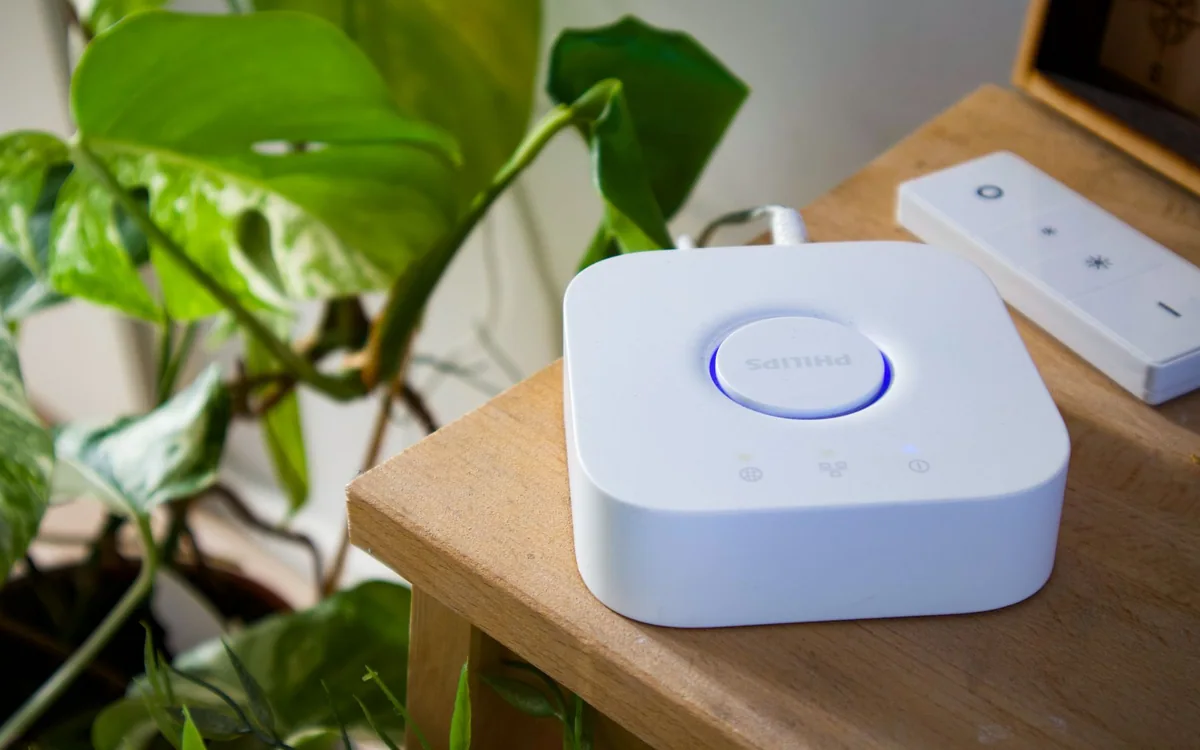 A white smart home hub beside a leafy plant on a wooden table.