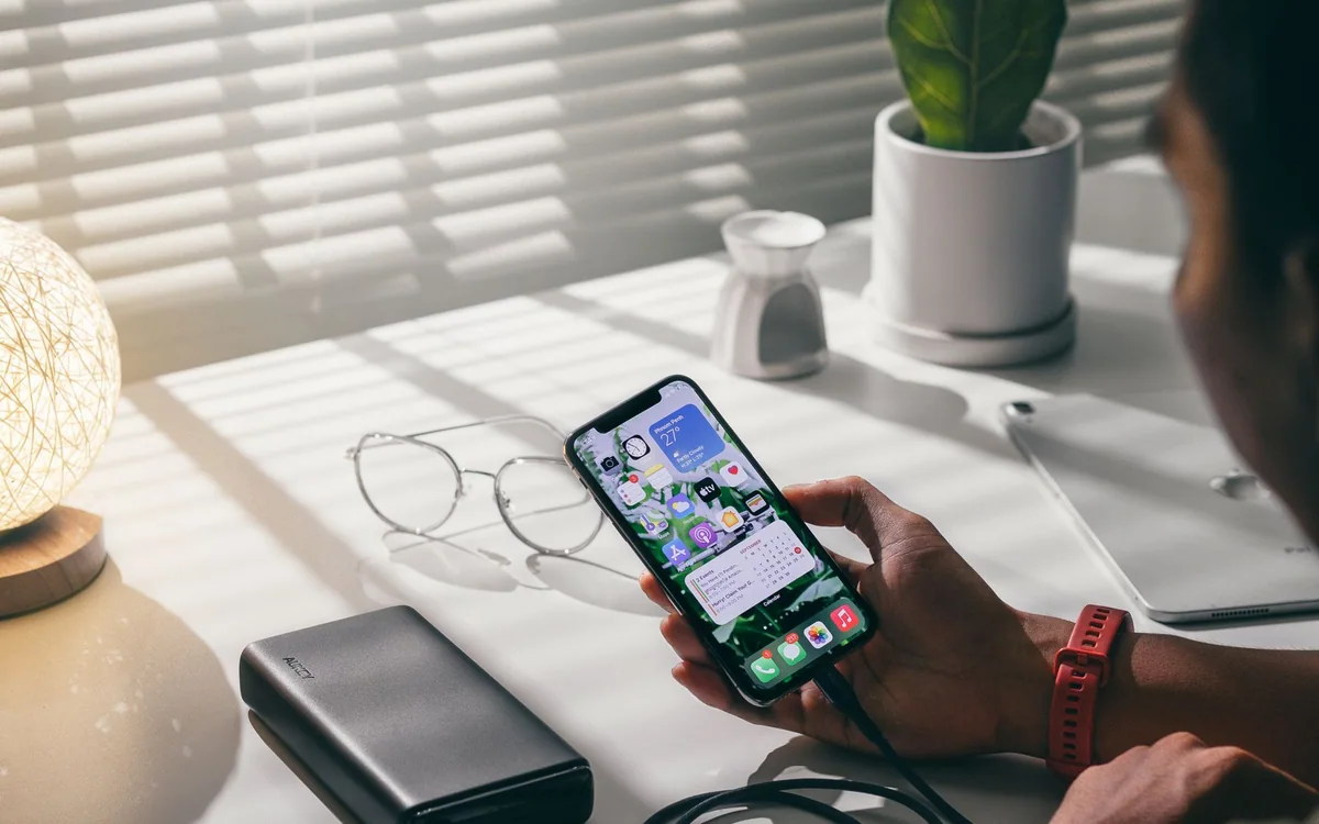 A hand holds a smartphone connected to a power bank on a desk.