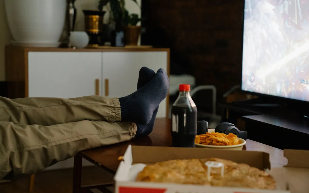 A person relaxes in front of a television with headphones and snacks on the table.