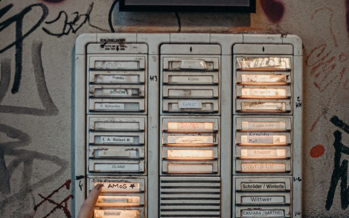 A hand presses an apartment entrance intercom mounted on a graffiti-covered wall.